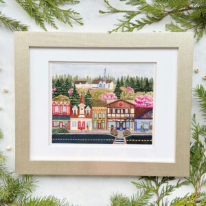 Whimsical painting of downtown Poulsbo WA during the spring with colorful shops, evergreen and blossoming cherry trees, and a white and blue church in a gold frame.